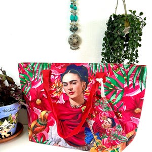 Könnte beinhalten: Eine Tragetasche mit einem lebendigen Blumendesign und einem Porträt von Frida Kahlo. Die Tasche hat rote Griffe und ein passendes Etikett. Der Hintergrund ist eine Mischung aus rosa und grünen Blumenmustern, was ein farbenfrohes Accessoire ergibt.