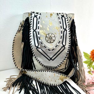 Può includere: Borsa a tracolla in pelle bianca e beige con frange nere e dettagli dorati. La borsa presenta una patta decorativa con cuciture nere e un abbellimento metallico. Anche la tracolla è in pelle e ha frange.