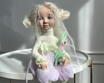 May include: A handmade doll with a serene expression, wearing a cream-colored sweater with ruffled sleeves, lavender bloomers, and green leggings. The doll has white hair styled in buns and holds a green crocheted bunny. It is seated on a white table with gold legs.