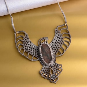 May include: A gray macrame necklace with a brown stone pendant in the shape of an eagle. The necklace is decorated with gold beads.