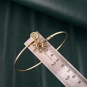Puede incluir: Un brazalete dorado con un dije en forma de sol con una piedra de ojo de tigre. El brazalete se mide en una regla marcada en pulgadas.