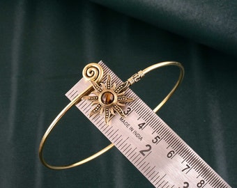 May include: A gold-toned bangle bracelet with a sun-shaped charm featuring a tiger's eye stone. The bracelet is measured on a ruler marked in inches.