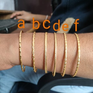 Può includere: Sei braccialetti d'oro con diversi motivi incisi. I braccialetti sono impilati sul polso di una persona. I braccialetti sono etichettati con a, b, c, d, e ed f.