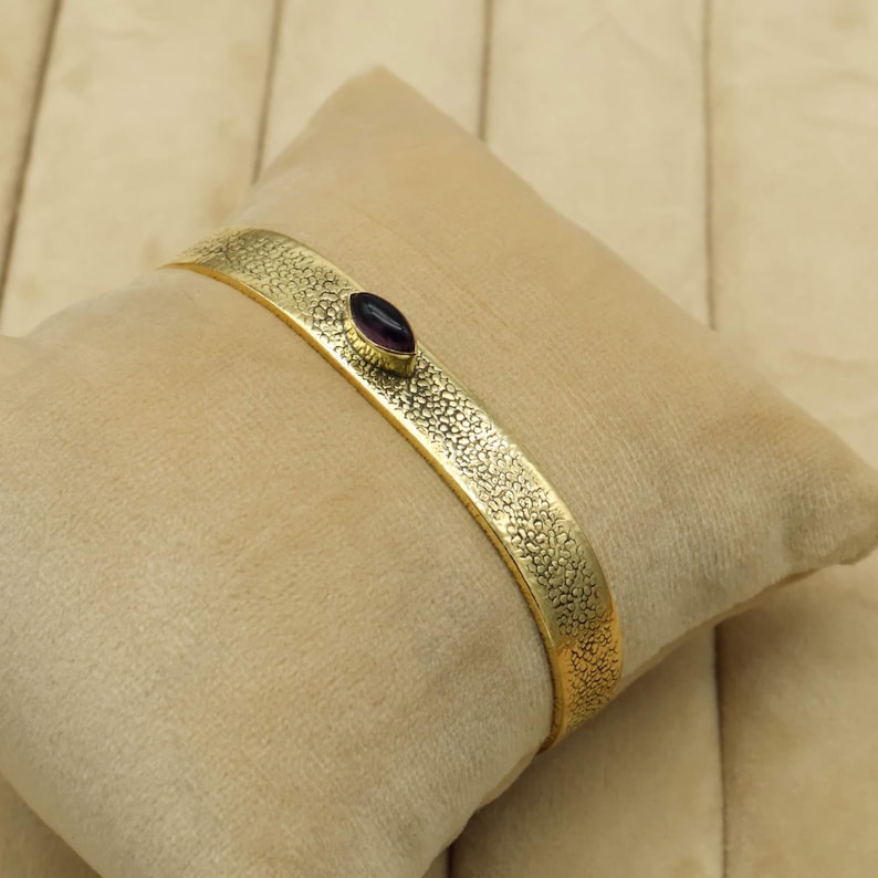 May include: A gold-colored cuff bracelet with a textured surface and a dark purple oval-shaped stone. The bracelet is resting on a beige pillow, showcasing its design and craftsmanship. This piece is a stylish accessory.