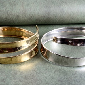 Pode incluir: Dois braceletes de metal martelado, um dourado e outro prateado, são exibidos em uma superfície verde texturizada. Os braceletes estão abertos e têm uma forma ligeiramente curva.