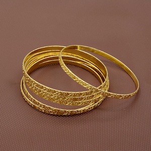 May include: A set of six gold-toned bangles with a delicate, embossed floral pattern. The bangles are stacked together and arranged on a brown surface.
