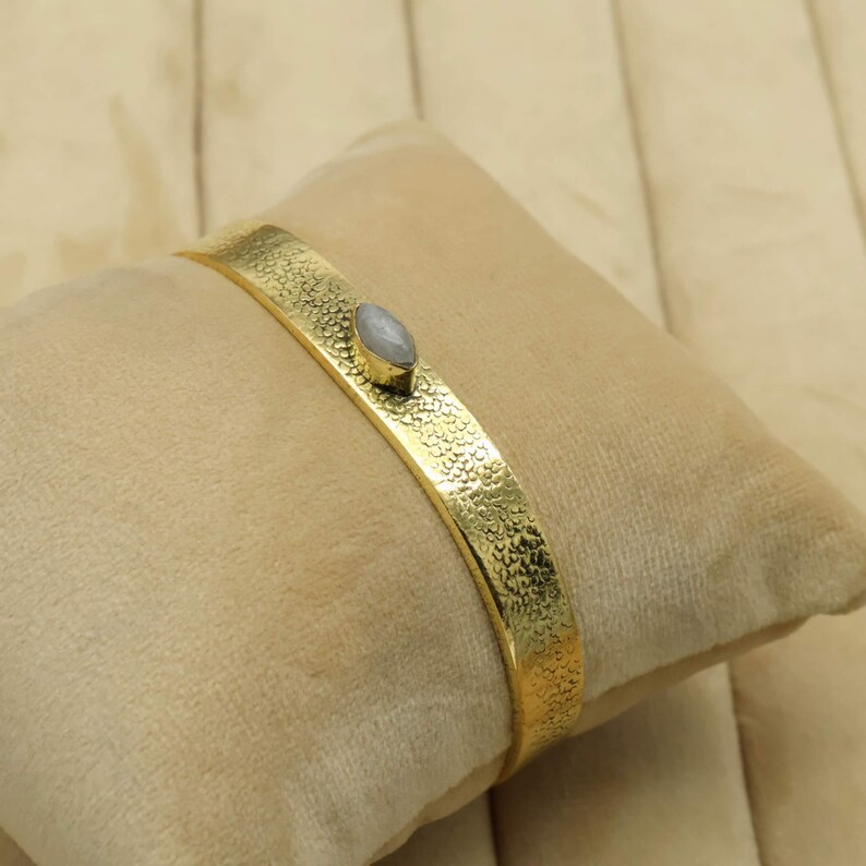 May include: A gold-colored cuff bracelet with a textured surface and a small, oval-shaped, light-colored stone. The bracelet is resting on a beige, velvet-like surface.