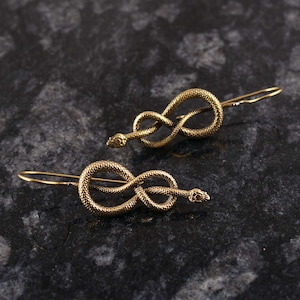 May include: A pair of gold-colored snake earrings. Each earring features a coiled snake design with a detailed head and tail, and a simple hook for pierced ears. The earrings are displayed on a dark, textured surface.