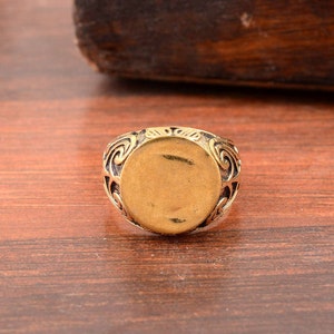 Puede incluir: Un anillo de tono dorado con un centro ovalado y detalles intrincados de volutas alrededor de los bordes.