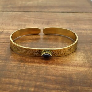 May include: A gold-colored cuff bracelet with a textured surface and a small, oval, dark blue stone. The bracelet is open at the ends and sits on a wooden surface.