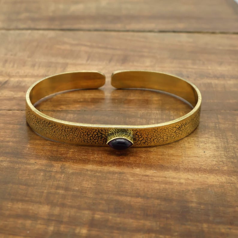 May include: A gold-colored cuff bracelet with a textured surface and a small, oval, dark blue stone. The bracelet is open at the ends and sits on a wooden surface.