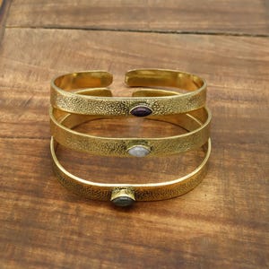 May include: Three gold-colored metal cuff bracelets with textured surfaces. Each bracelet features a small, oval-shaped gemstone: purple, white, and green. The bracelets are stacked on a wooden surface.