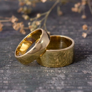 May include: Two gold wedding bands, one with a smooth finish and the other with a textured finish, are stacked on top of each other on a wooden surface.