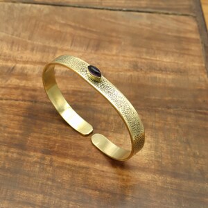 May include: A gold-colored cuff bracelet with a textured surface and a small, oval, dark purple stone. The open-ended bracelet is displayed on a wooden surface, showcasing its design and craftsmanship.