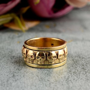 May include: A gold-toned ring with a band of skulls around the circumference. The skulls are detailed and have a slightly raised design.