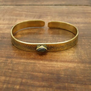 May include: A gold-colored cuff bracelet with a textured surface and a small, dark-colored stone set in the center. The open-ended design allows for easy wear. The bracelet is displayed on a wooden surface.