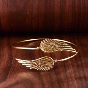 May include: A gold-toned bangle bracelet featuring two wing-shaped charms. The wings are intricately detailed with a textured design.