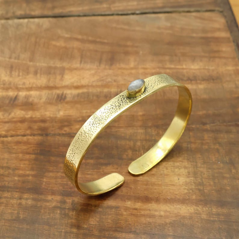 May include: A gold-colored cuff bracelet with a textured surface and an oval, light-colored stone set in the center. The open-ended bracelet is displayed on a wooden surface.