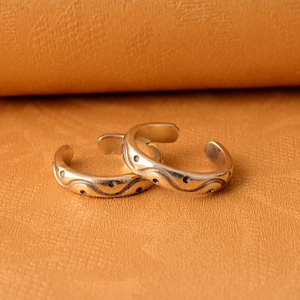 Può includere: Due anelli per dita dei piedi in argento con un design ondulato e piccoli punti. Gli anelli sono su una superficie in pelle marrone.