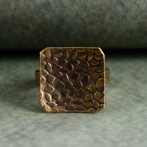 Peut inclure: Une bague carrée en laiton avec une surface texturée. La surface est recouverte d'un motif de petits cercles et ovales en relief.