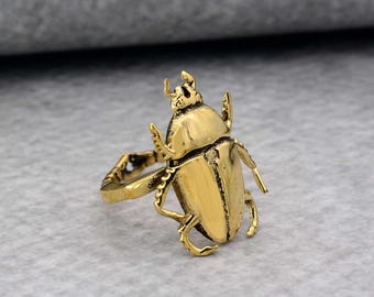 May include: A gold-toned ring featuring a detailed beetle design. The ring is made of metal and has a textured finish.