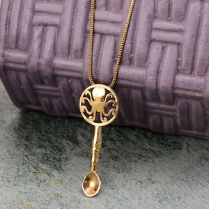 Puede incluir: Un collar de oro con un colgante con forma de cuchara con un diseño de pulpo. El pulpo está recortado en la parte superior redonda de la cuchara.