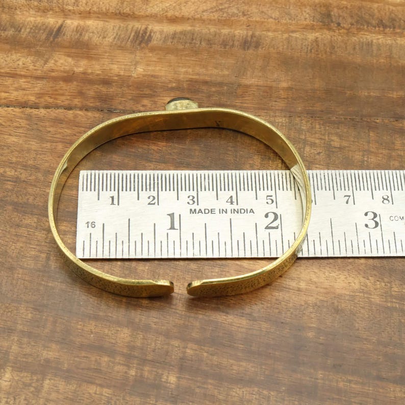 May include: A gold-colored metal bracelet with a small, dark stone set in the center. The bracelet is open-ended and rests on a wooden surface next to a ruler. The ruler shows measurements in inches.