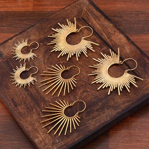 May include: Set of four gold-toned hoop earrings with a sunburst design. The earrings are of varying sizes and are displayed on a wooden surface.