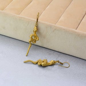 Peut inclure: Boucles d'oreilles serpent dorées. Les boucles d'oreilles présentent un motif de serpent détaillé avec un corps enroulé et une queue texturée. Les boucles d'oreilles ont une fermeture à crochet et sont présentées sur une surface gris clair.