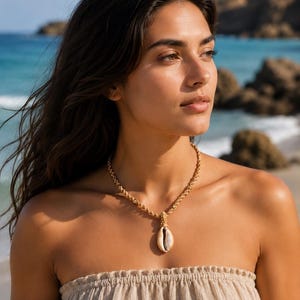 May include: A gold-toned rope necklace with a seashell pendant. The necklace is worn by a person wearing a beige strapless top. The background shows a beach with blue water and rocks.