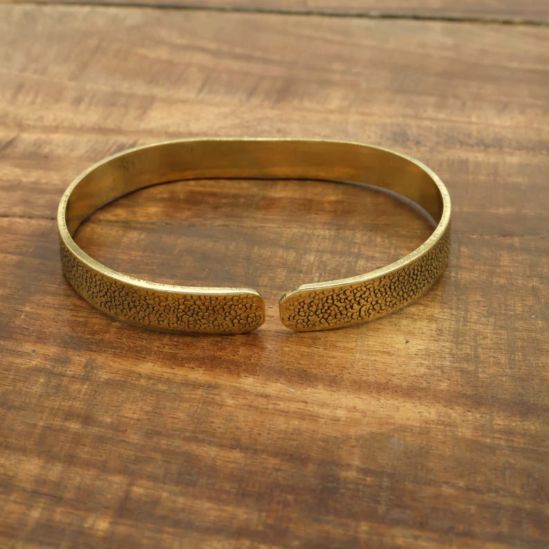 May include: A gold-colored cuff bracelet with a textured surface. The open-ended bracelet is oval-shaped and rests on a wooden surface. The bracelet's surface has a speckled pattern.