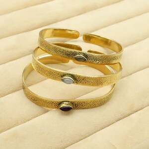 May include: Three gold-colored cuff bracelets with textured surfaces and small, oval-shaped gemstones. The gemstones are in shades of gray and purple. The bracelets are stacked on top of each other, with the top one open.