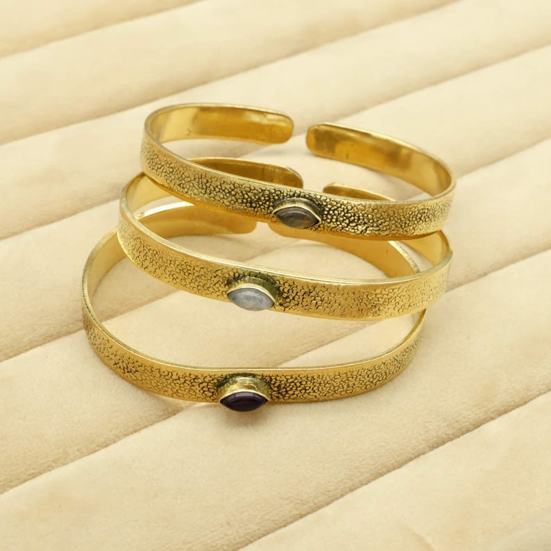 May include: Three gold-colored cuff bracelets with textured surfaces and small, oval-shaped gemstones. The gemstones are in shades of gray and purple. The bracelets are stacked on top of each other, with the top one open.