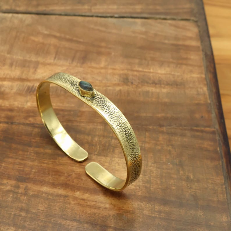 May include: A gold-colored cuff bracelet with a textured surface and a small, dark-colored stone set in the center. The bracelet is open-ended and rests on a wooden surface.