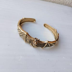 Op de afbeelding: Een gouden metalen armband met een vleermuisontwerp. De vleermuis bevindt zich in het midden van de armband en heeft gedetailleerde vleugels en een textuurrijk lichaam.