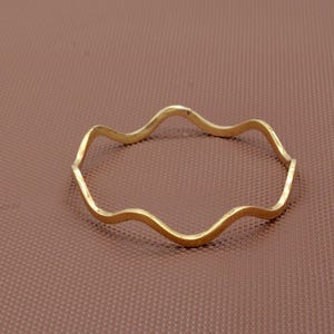 Peut inclure: Un bracelet jonc doré avec un design ondulé et irrégulier. Le bracelet a une forme continue et fluide, et est posé sur un fond marron. Le bracelet est un bijou.