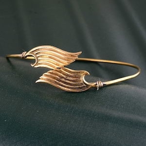 May include: A gold-toned bracelet featuring a wing design. The wings are detailed with lines and are connected to a thin, circular band.