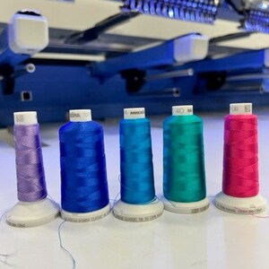 May include: Five spools of embroidery thread in different colors: purple, blue, teal, turquoise, and pink. The spools are arranged in a row on a white surface.