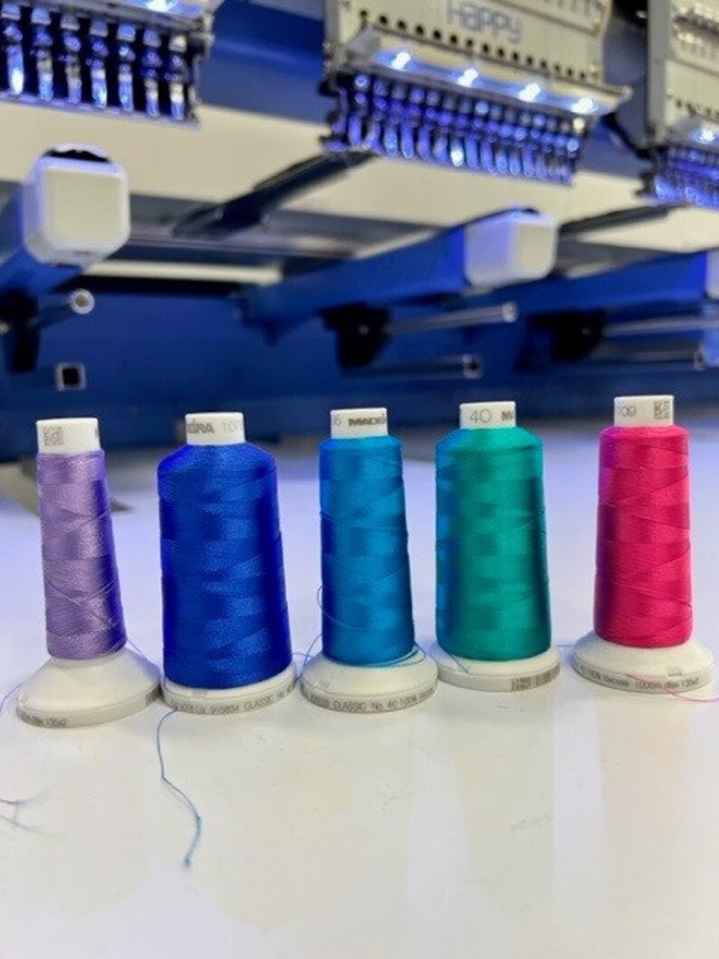 May include: Five spools of embroidery thread in different colors: purple, blue, teal, turquoise, and pink. The spools are arranged in a row on a white surface.