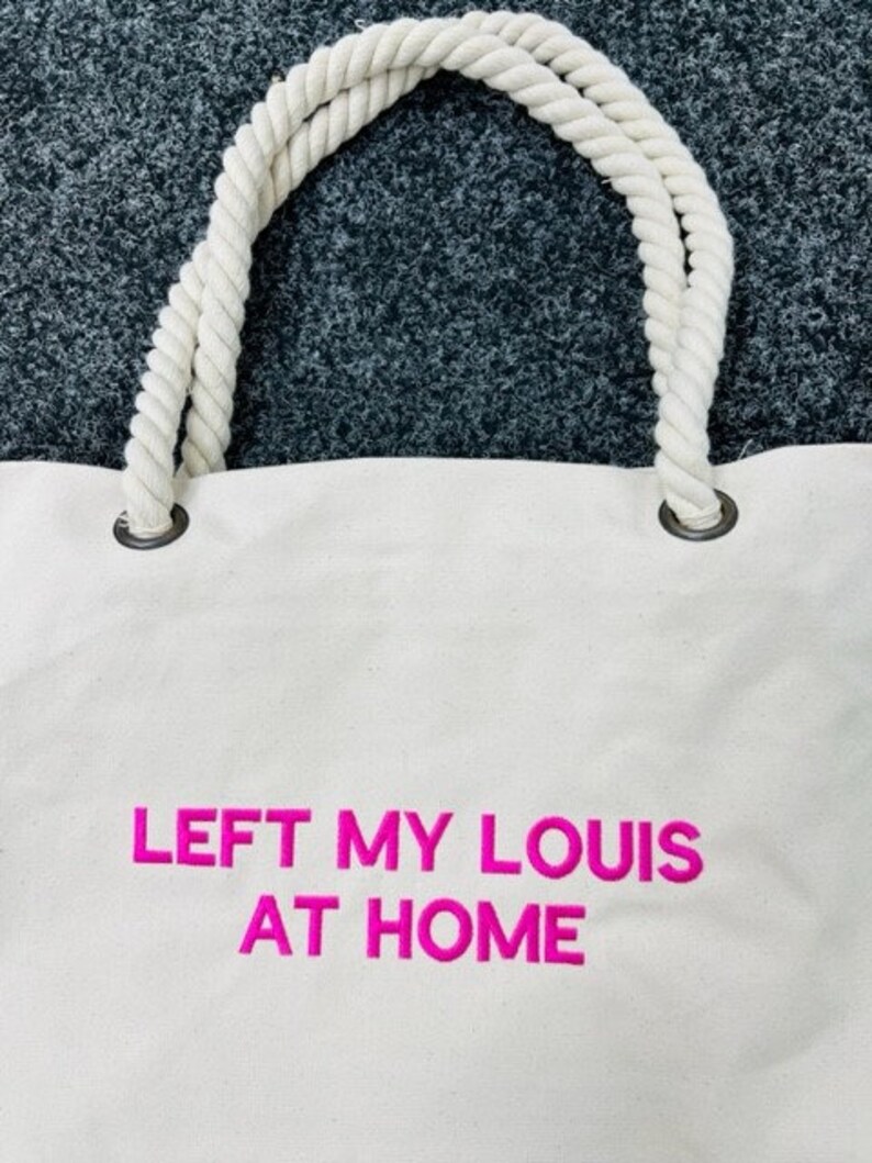 May include: White canvas tote bag with thick white rope handles. The bag has pink embroidered text that reads "LEFT MY LOUIS AT HOME".