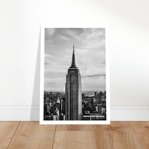 May include: A black and white photograph of the Empire State Building in New York City. The building is tall and slender, with a pointed spire at the top. The photo was taken from a high vantage point, looking down on the city.