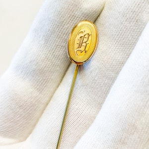 May include: A gold-toned stick pin with an oval-shaped head featuring an engraved letter "B".