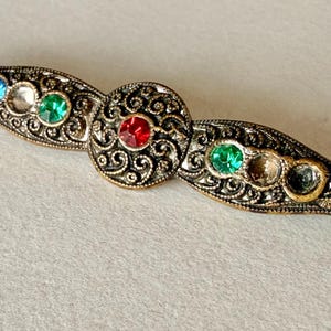 Könnte beinhalten: Eine lange, schmale Brosche aus Metall im Antik-Stil mit einem komplexen, erhabenen Schnörkelmuster. Das Stück verfügt über ein zentrales, rundes Medaillon mit einem roten Stein, flankiert von zwei grünen, einem klaren und einem blauen Stein.