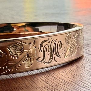 Puede incluir: Un brazalete rígido de metal en tono dorado con un intrincado patrón floral grabado y un monograma central con las letras D, S y M entrelazadas en una escritura decorativa. La superficie pulida refleja la luz en su exterior curvo.