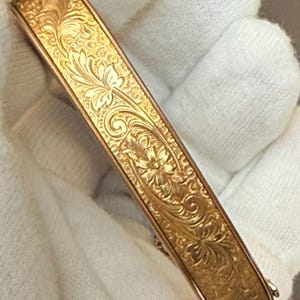 Op de afbeelding: Een goudkleurige scharnierende armband met een ingewikkeld, in reliëf aangebracht bloemen- en krulpatroon over het gehele buitenoppervlak. Het metaal heeft een gepolijste afwerking met verdiepte details die de gegraveerde botanische motieven benadrukken.