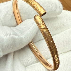 Puede incluir: Un brazalete de metal en tono dorado con un diseño abierto y un patrón floral en relieve continuo en la superficie exterior. El brazalete tiene un interior liso y pulido con un perfil redondeado, mostrando un acabado metálico brillante.