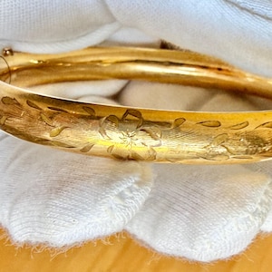May include: A polished gold bangle bracelet featuring a rounded profile and a delicate, hand-engraved floral pattern that wraps around the exterior surface. The bracelet includes a secure hinged clasp mechanism visible on the side of the band.