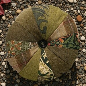 May include: A circular cushion featuring a patchwork design with a central black button. The cushion is made from various green and brown fabrics, including floral, plaid, and textured patterns. The design incorporates a variety of textures and shades.