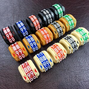 Könnte beinhalten: Eine Sammlung von bunten, nummerierten Kunststoffringen. Die Ringe sind in verschiedenen Farben, darunter Rot, Blau, Grün, Schwarz und Weiß. Jeder Ring hat eine darauf gedruckte Nummer, wobei die Zahlen von 77 bis 88 reichen. Die Ringe sind in einem Halbkreis auf einer braunen Oberfläche angeordnet.