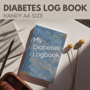 May include: A blue and gold marbled journal with the text "My Diabetes Logbook." The journal is on a white surface with two yellow dried flowers.